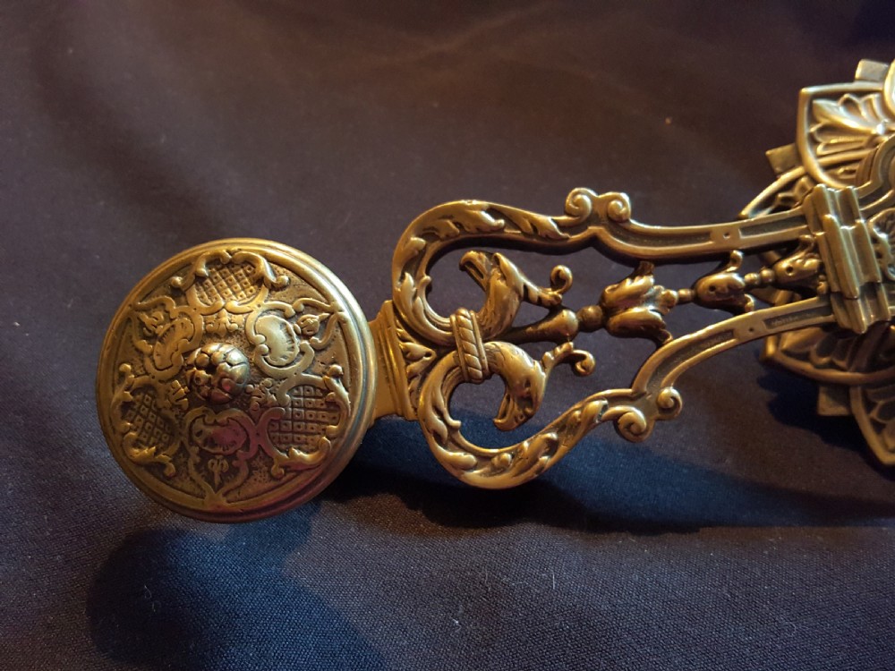 An ornate cast brass mechanical wall-mounted bell ringer featuring a sunburst rosette top, openwork scrollwork with mythical creatures, and a circular decorative pendulum drop. View VI