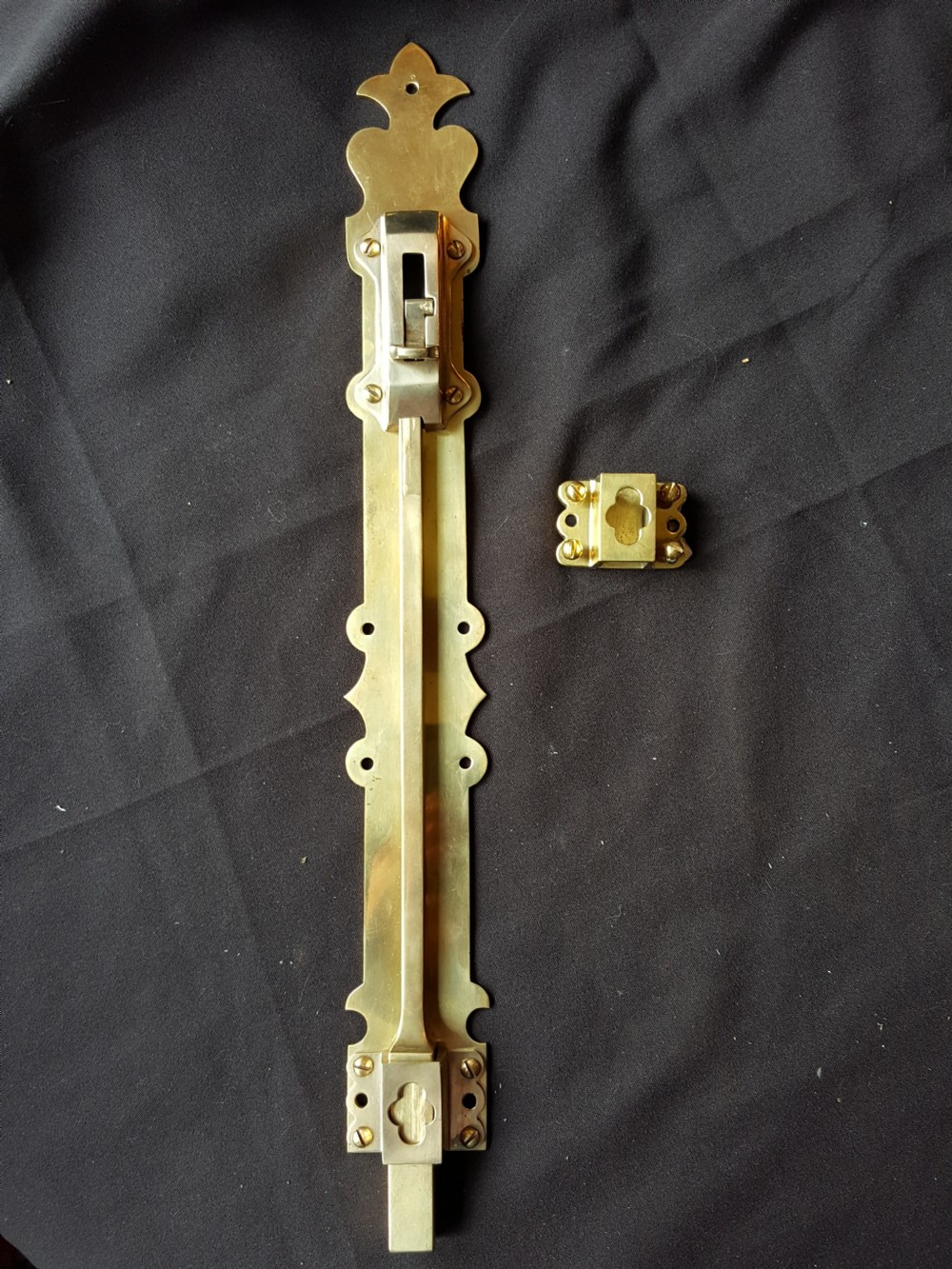 A Victorian Gothic Revival solid brass surface door bolt with a decorative shaped backplate, trefoil cutouts, and a matching keeper.