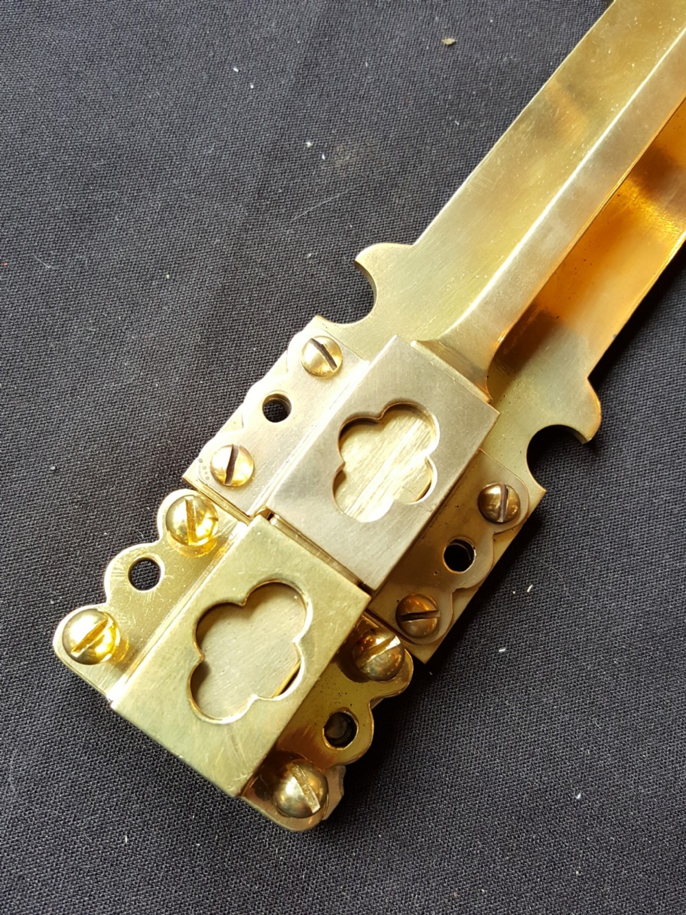 A Victorian Gothic Revival solid brass surface door bolt with a decorative shaped backplate, trefoil cutouts, and a matching keeper. View IV