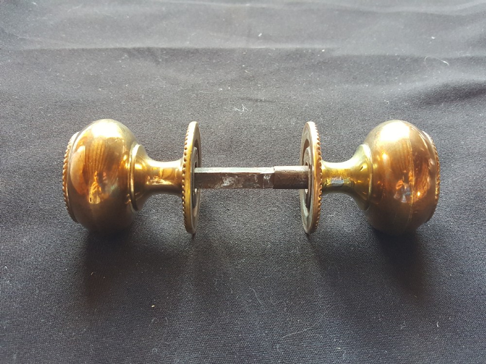 A pair of polished Regency brass door knobs joined by a square spindle, featuring beaded rims and central floral relief details. View IV