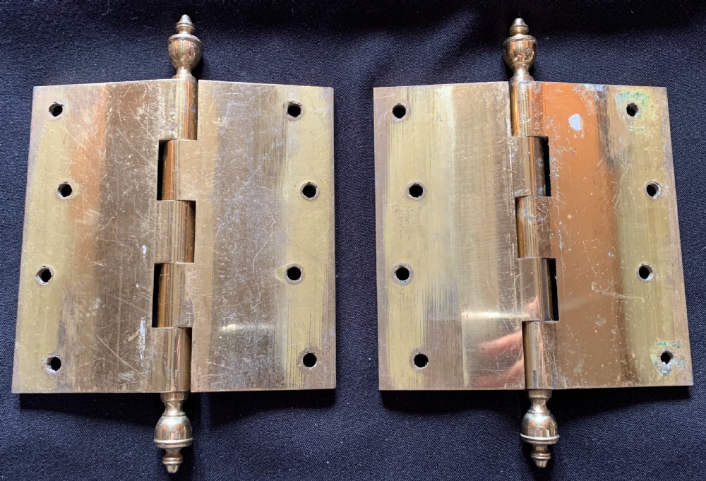 A pair of solid brass Victorian butt door hinges featuring turned urn finials at the top and bottom of each hinge pin.