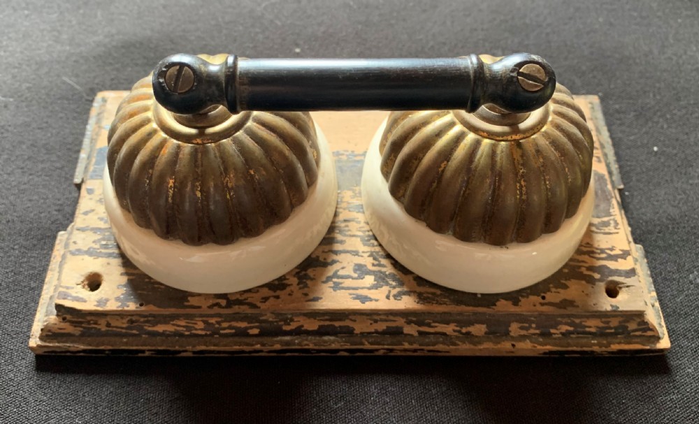 An antique Victorian double lever light switch featuring fluted brass domes on white porcelain bases, connected by a dark wooden lever bar on a timber backplate.