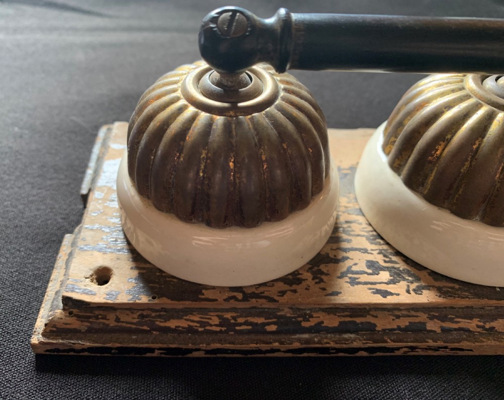 An antique Victorian double lever light switch featuring fluted brass domes on white porcelain bases, connected by a dark wooden lever bar on a timber backplate. View III