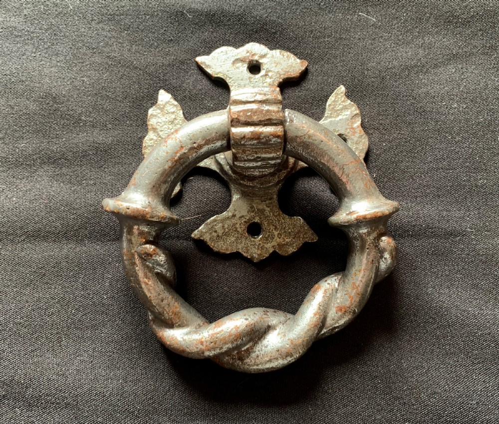 A dark cast iron door knocker featuring a twisted ring handle suspended from a decorative gothic mounting plate.