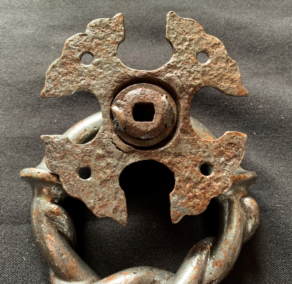 A dark cast iron door knocker featuring a twisted ring handle suspended from a decorative gothic mounting plate. View V