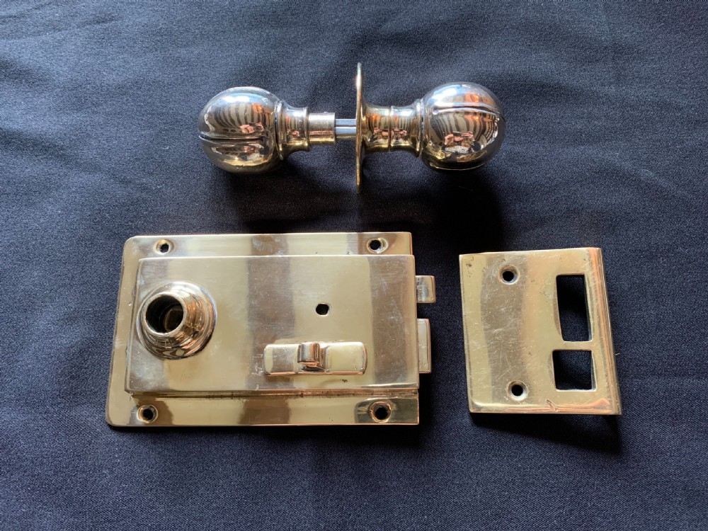 A polished brass Art Nouveau rim lock set featuring a rectangular surface lock, matching keep plate, and a pair of round fluted brass door knobs on a spindle.