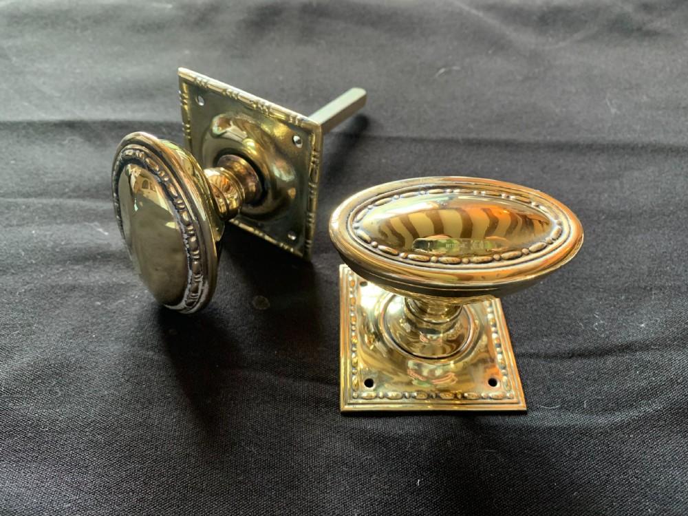 A pair of polished oval brass door knobs featuring beaded rims mounted on square decorative backplates with a connecting square metal spindle.