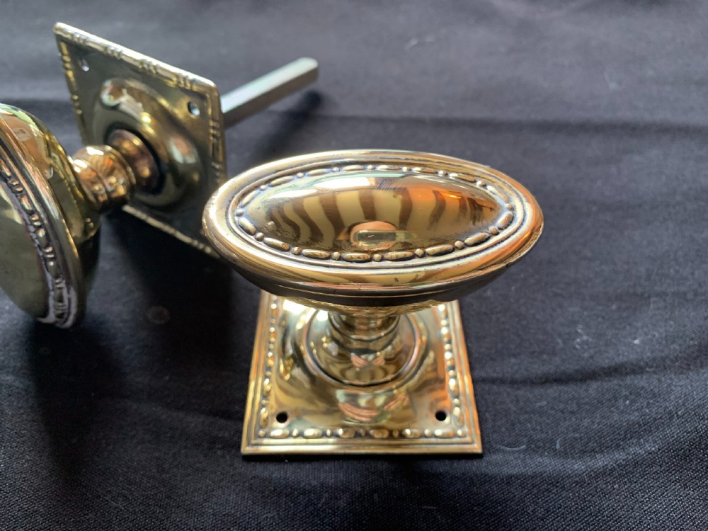A pair of polished oval brass door knobs featuring beaded rims mounted on square decorative backplates with a connecting square metal spindle. View II