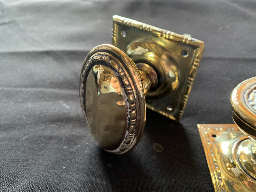 A pair of polished oval brass door knobs featuring beaded rims mounted on square decorative backplates with a connecting square metal spindle. View III
