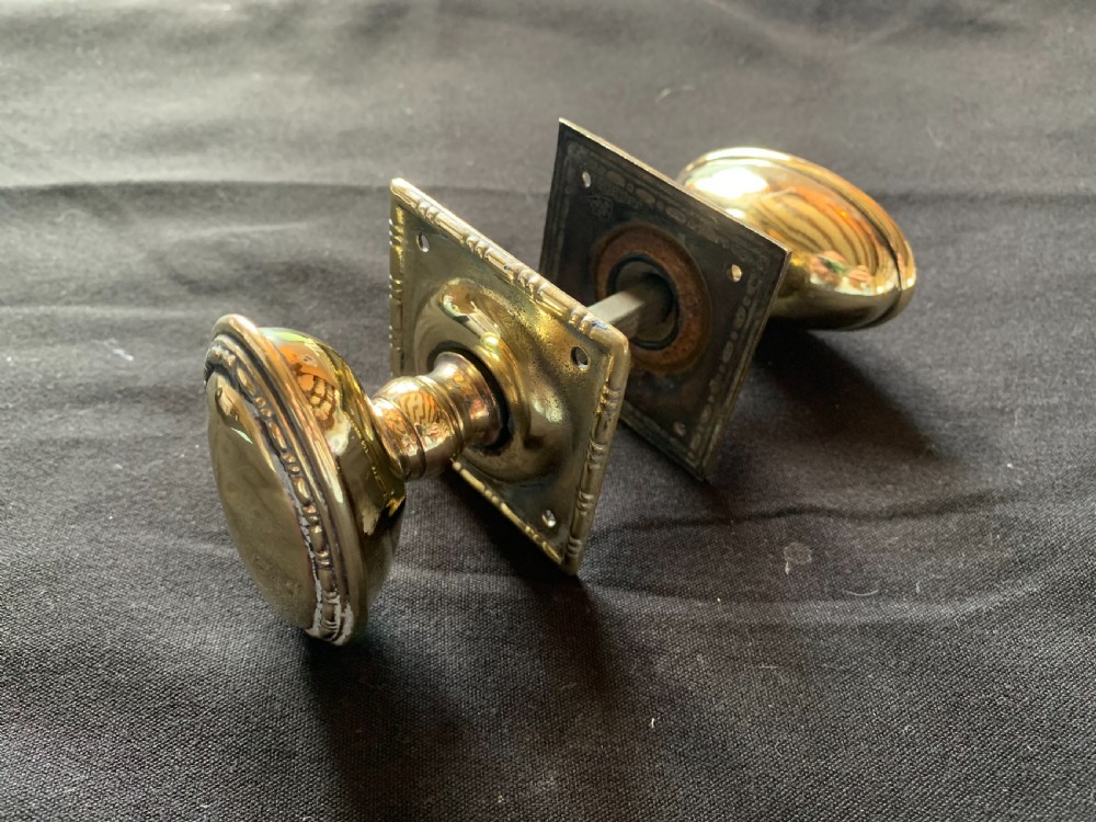 A pair of polished oval brass door knobs featuring beaded rims mounted on square decorative backplates with a connecting square metal spindle. View IV