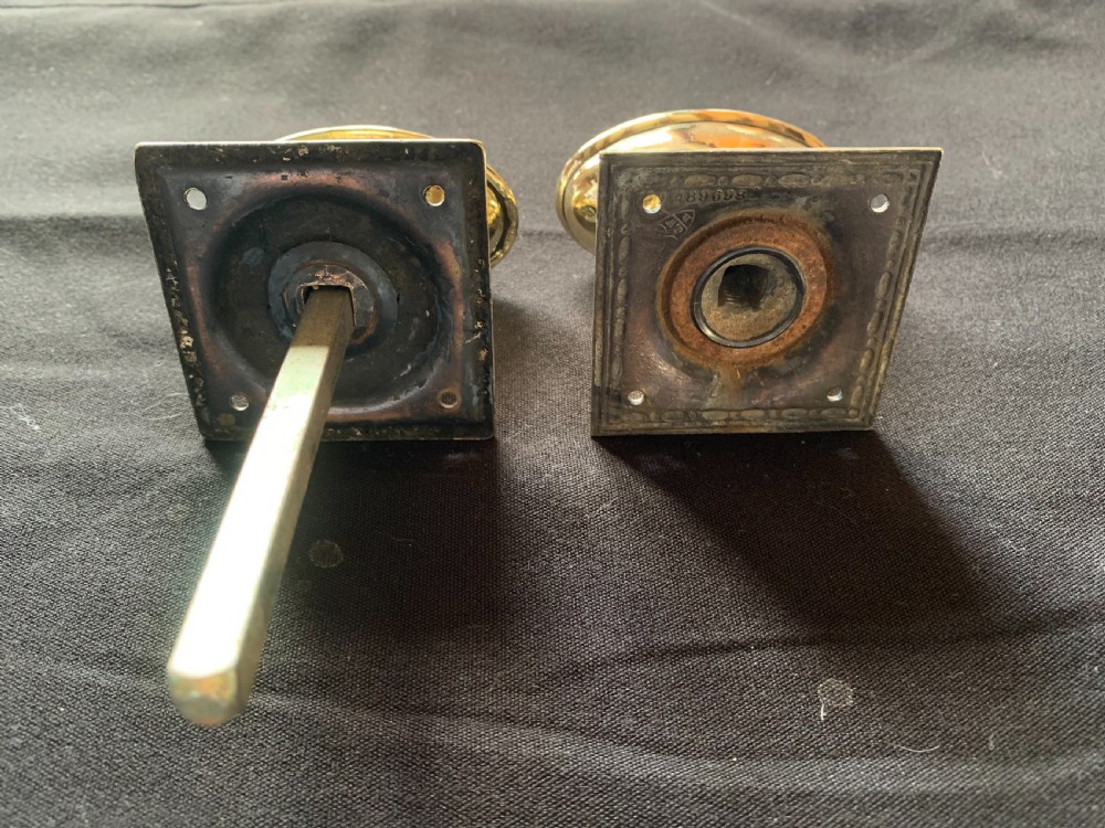 A pair of polished oval brass door knobs featuring beaded rims mounted on square decorative backplates with a connecting square metal spindle. View V