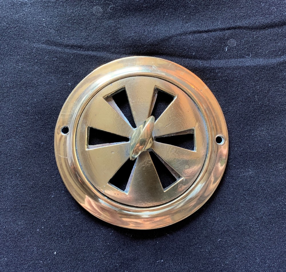 A circular solid brass Victorian hit-and-miss air vent with a central turning handle, triangular slotted openings, and two mounting screw holes.