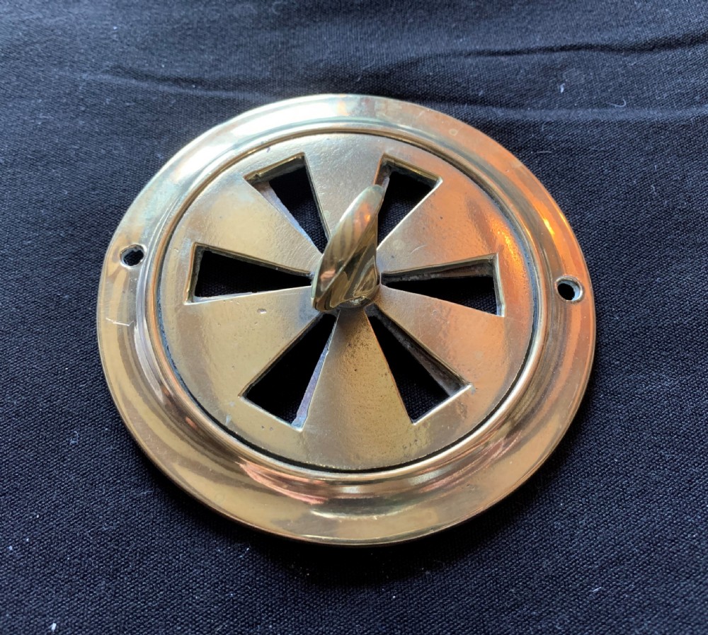 A circular solid brass Victorian hit-and-miss air vent with a central turning handle, triangular slotted openings, and two mounting screw holes. View II