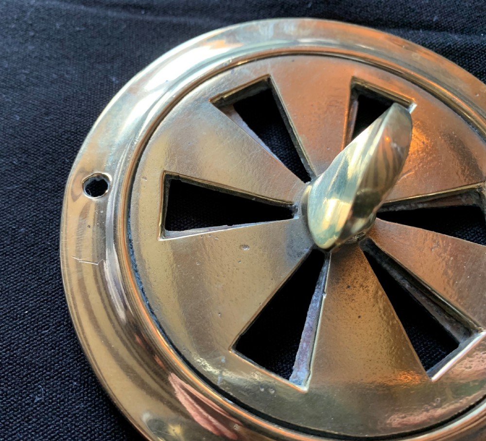 A circular solid brass Victorian hit-and-miss air vent with a central turning handle, triangular slotted openings, and two mounting screw holes. View III