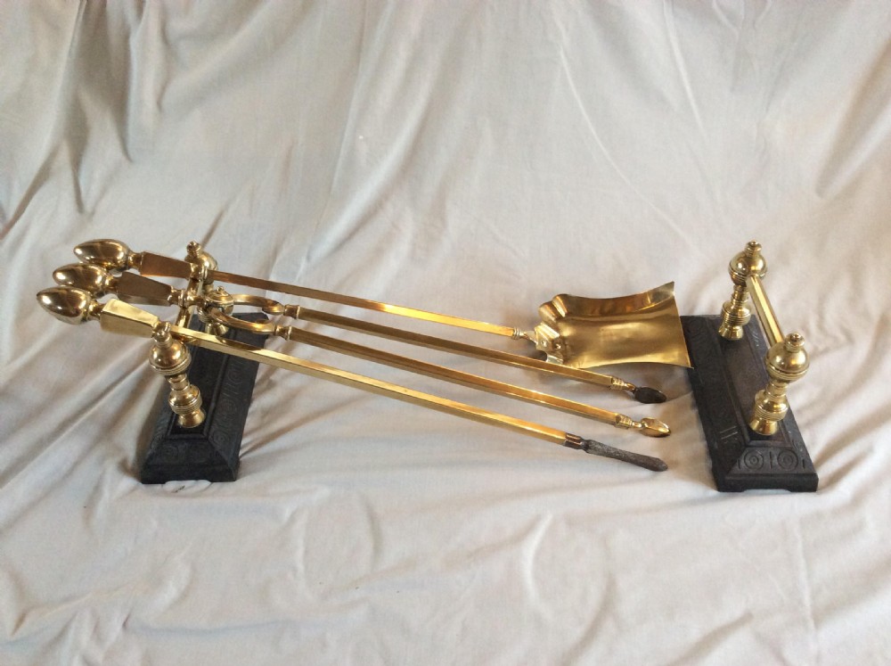 An antique Arts and Crafts companion set featuring brass fire tools including a shovel, tongs, and poker, supported on black cast iron and brass rests.