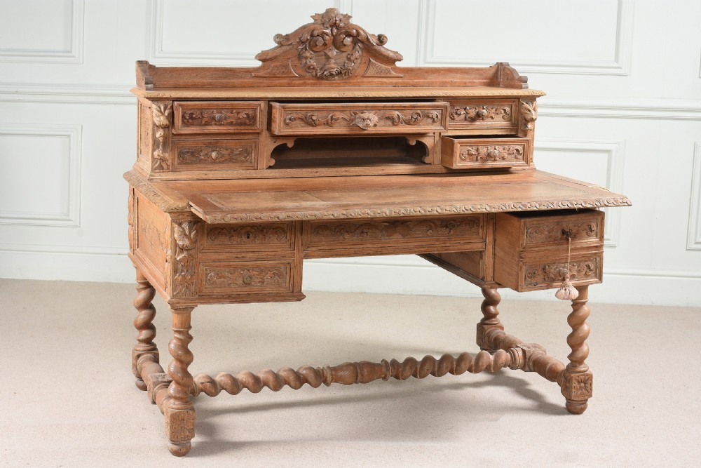 A carved oak writing desk featuring a top shelf with small drawers, an ornate crest, a pull-out writing surface, and barley twist legs connected by stretchers.