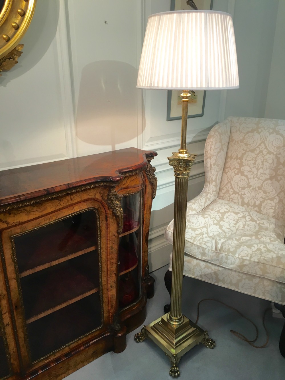 A brass telescopic standard lamp featuring a fluted column stem, Corinthian capital detail, a square stepped base with ornate feet, and a pleated white shade.