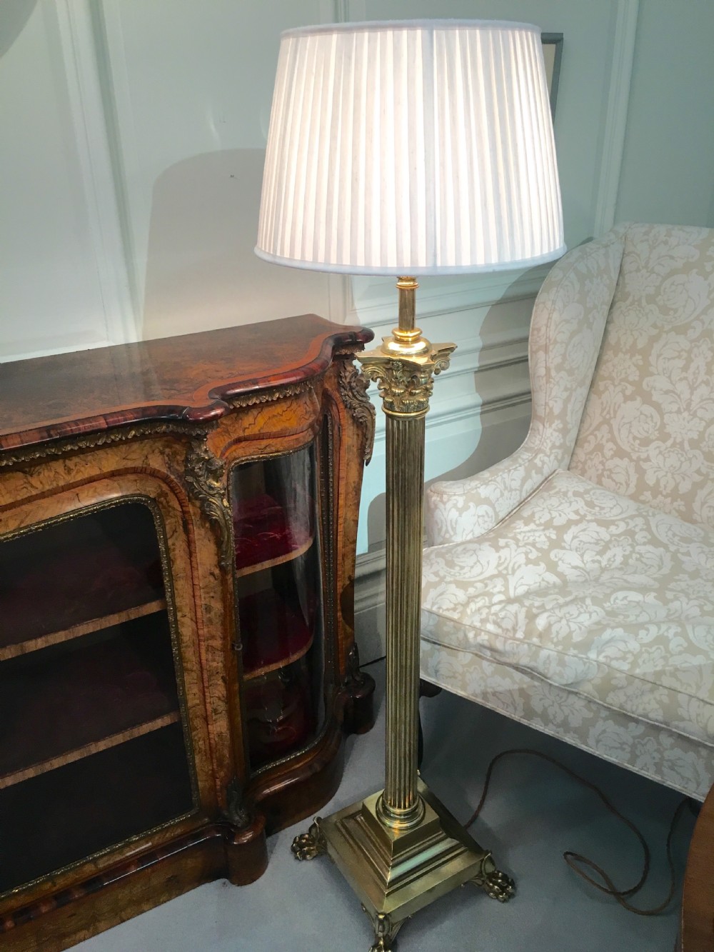 A brass telescopic standard lamp featuring a fluted column stem, Corinthian capital detail, a square stepped base with ornate feet, and a pleated white shade. View X