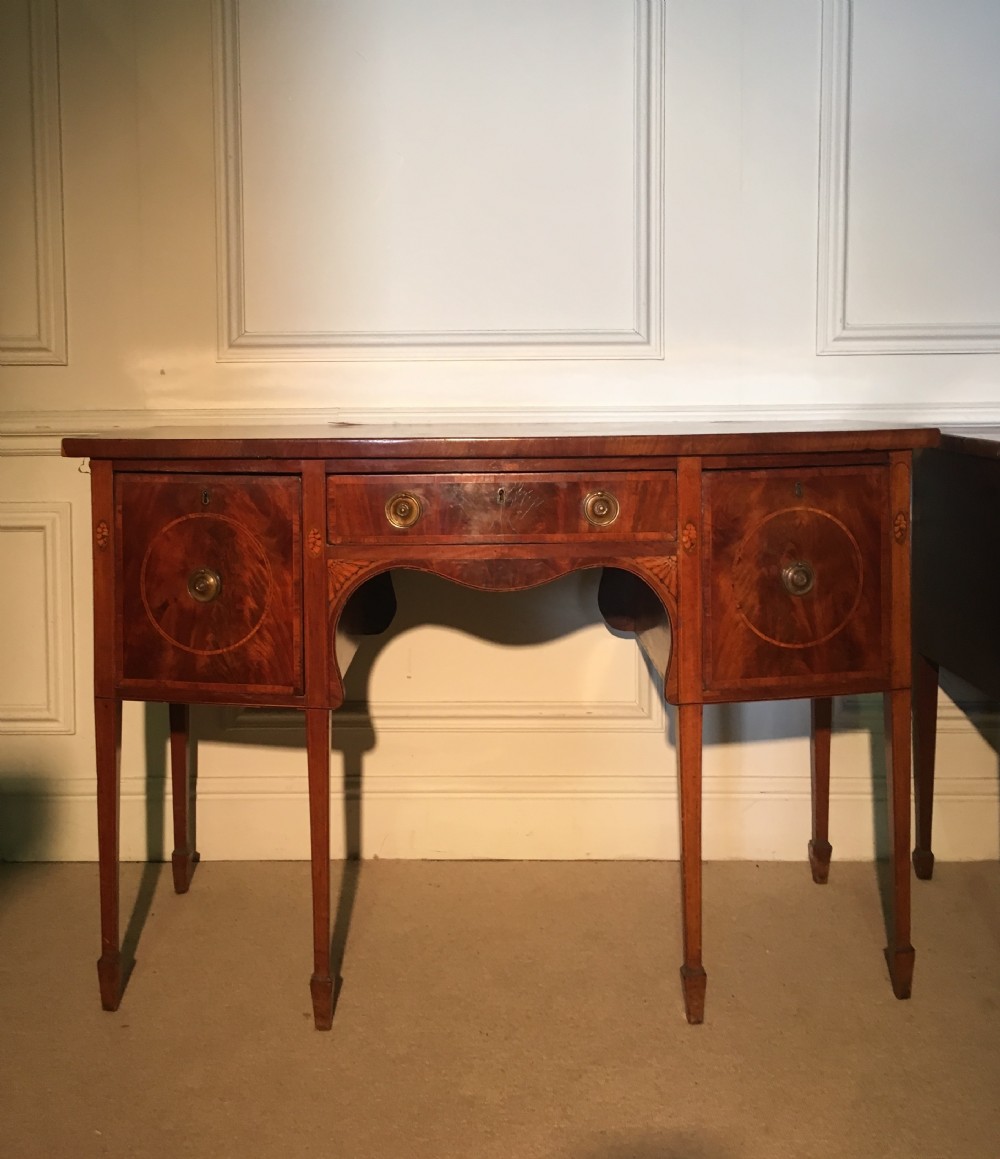 An antique mahogany Sheraton sideboard featuring satinwood inlay, square tapered legs, brass ring handles, and an arched central apron.