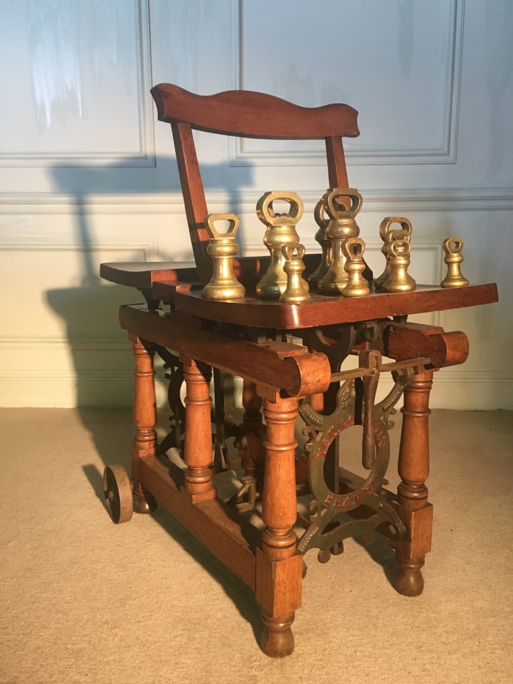 An antique oak jockey scale chair by Young & Son featuring turned legs, rear wheels, a brass balance mechanism, and a set of bell-shaped brass weights.
