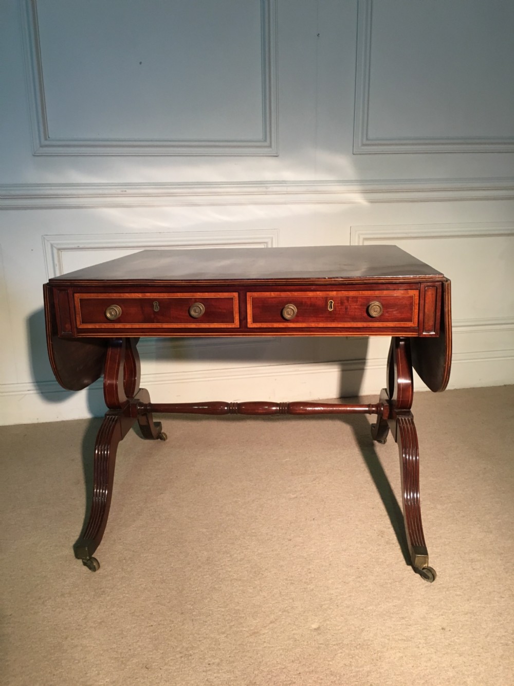 A George III Sheraton mahogany sofa table featuring two drop leaves, twin drawers with crossbanded inlay, lyre-form end supports, and reeded legs on brass castors. View II