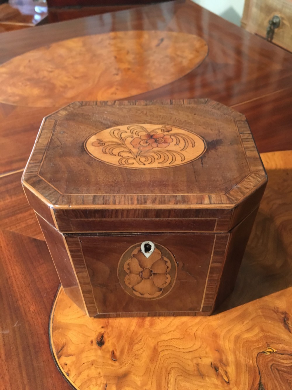 An octagonal George III wooden tea caddy featuring a hinged lid with a central floral marquetry medallion, crossbanded borders, and a matching front escutcheon inlay.