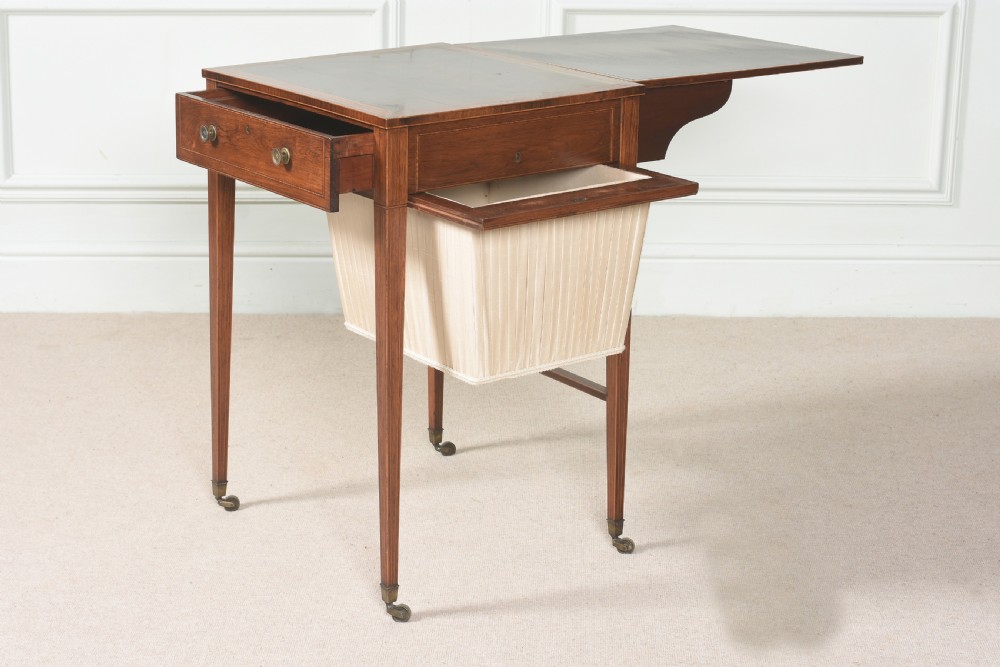 An 18th-century Sheraton rosewood sewing table featuring a top drawer, a suspended cream pleated fabric work bag, square tapered legs, and brass castors. View VI