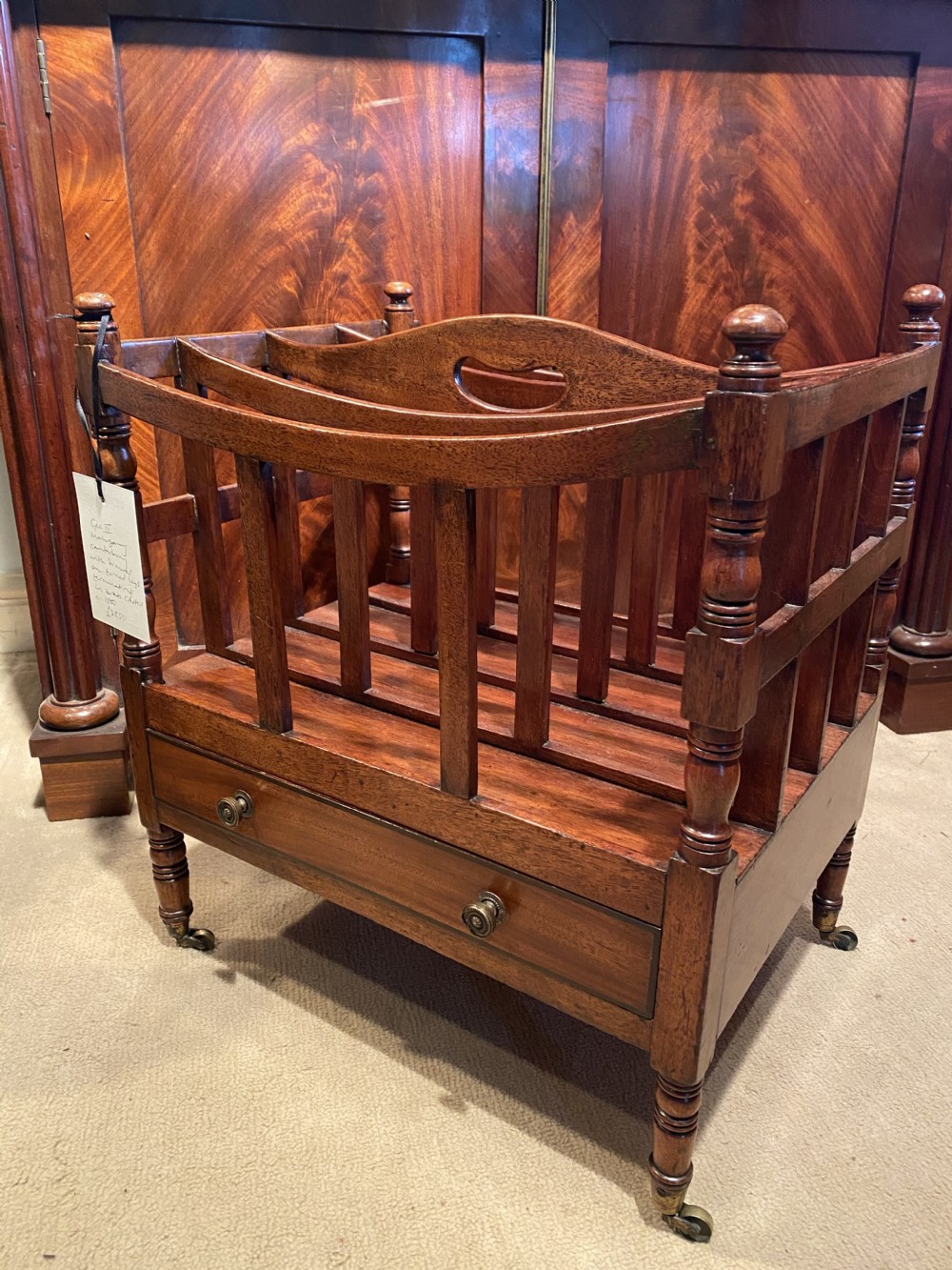 A George III mahogany canterbury featuring four slatted compartments, a single base drawer with brass knobs, and turned legs with brass castors.