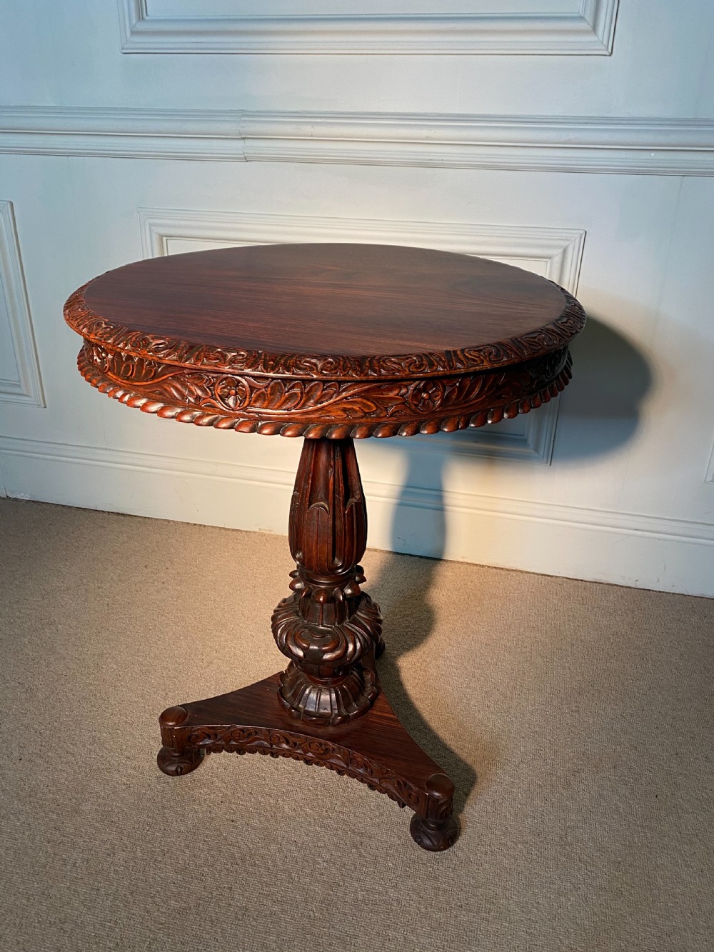 A circular Anglo-Indian padauk wood table featuring a heavily carved floral rim, turned central pedestal, and tripartite base.