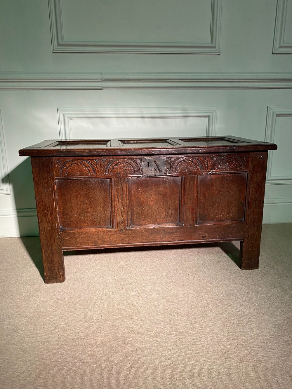 An 18th-century dark oak coffer with a carved upper frieze, three recessed front panels, iron lock plate, and a panelled top.