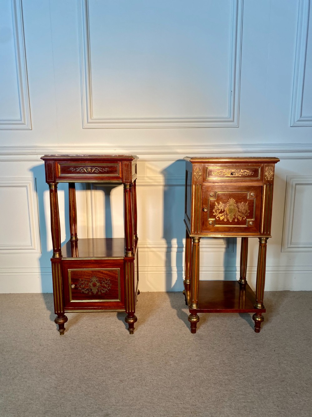 A pair of French Empire mahogany bedside cabinets featuring marble tops, inlaid brass classical foliage motifs, fluted column supports, and single cupboard doors.
