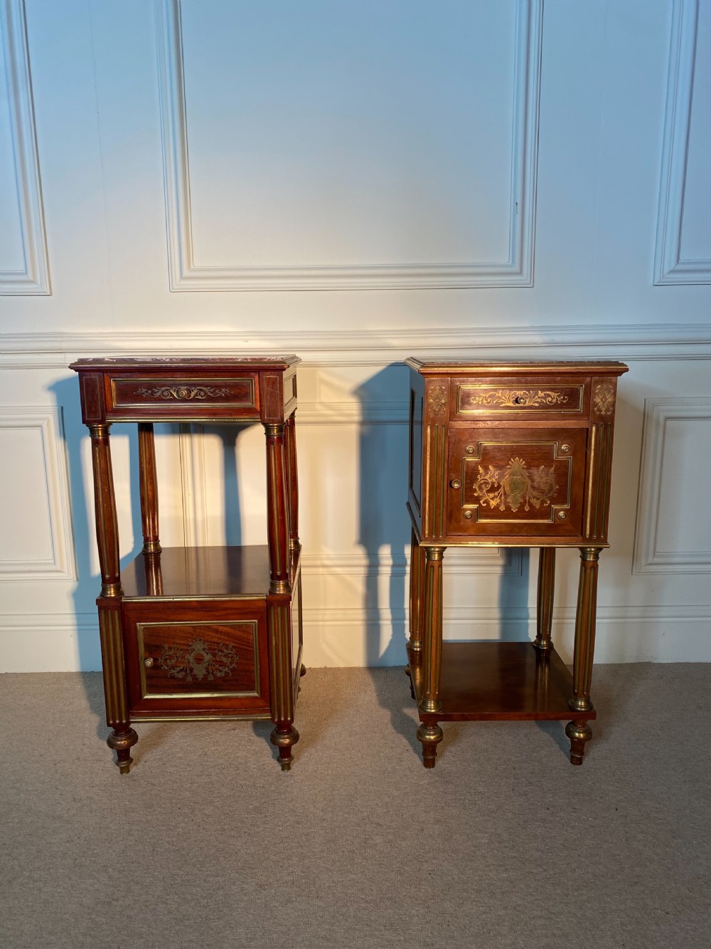 A pair of French Empire mahogany bedside cabinets featuring marble tops, inlaid brass classical foliage motifs, fluted column supports, and single cupboard doors. View II