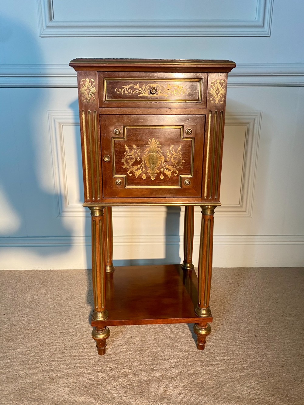 A pair of French Empire mahogany bedside cabinets featuring marble tops, inlaid brass classical foliage motifs, fluted column supports, and single cupboard doors. View IV