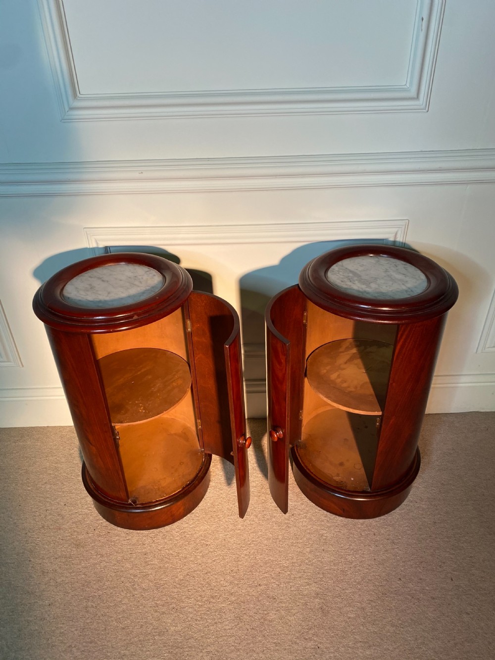 A pair of 19th-century cylindrical flame mahogany bedside cupboards featuring inset marble tops and single curved doors with turned wooden knobs. View VI