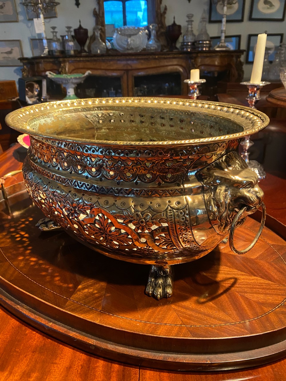 An oval polished brass wine cooler featuring intricate pierced repousse detailing, side lion mask handles with ring pulls, and four claw feet.