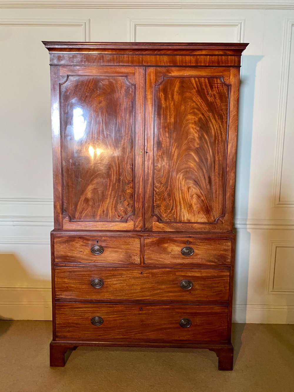 A George III mahogany linen press featuring flame mahogany panels on the upper cupboard doors above four drawers with brass handles and bracket feet. View III
