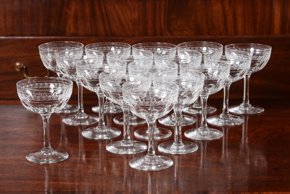 A set of sixteen clear etched crystal champagne coupes with diamond patterned rims displayed on a dark wood surface. View VII