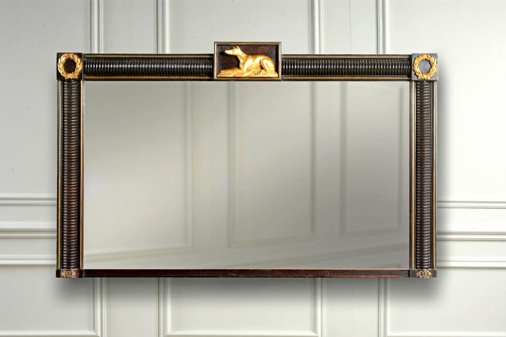 A rectangular Regency overmantel mirror featuring an ebonised reeded frame with gold laurel wreath corner ornaments and a central gilded greyhound motif at the top.