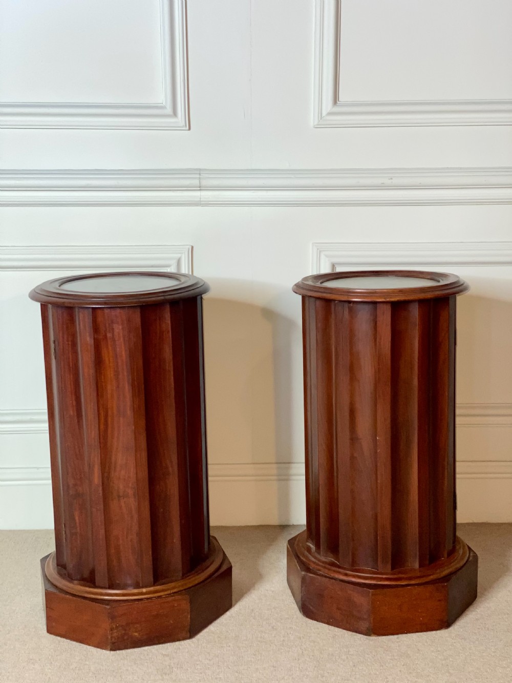 A pair of cylindrical mahogany bedside stands featuring fluted wooden columns, inset marble tops, and octagonal plinth bases.