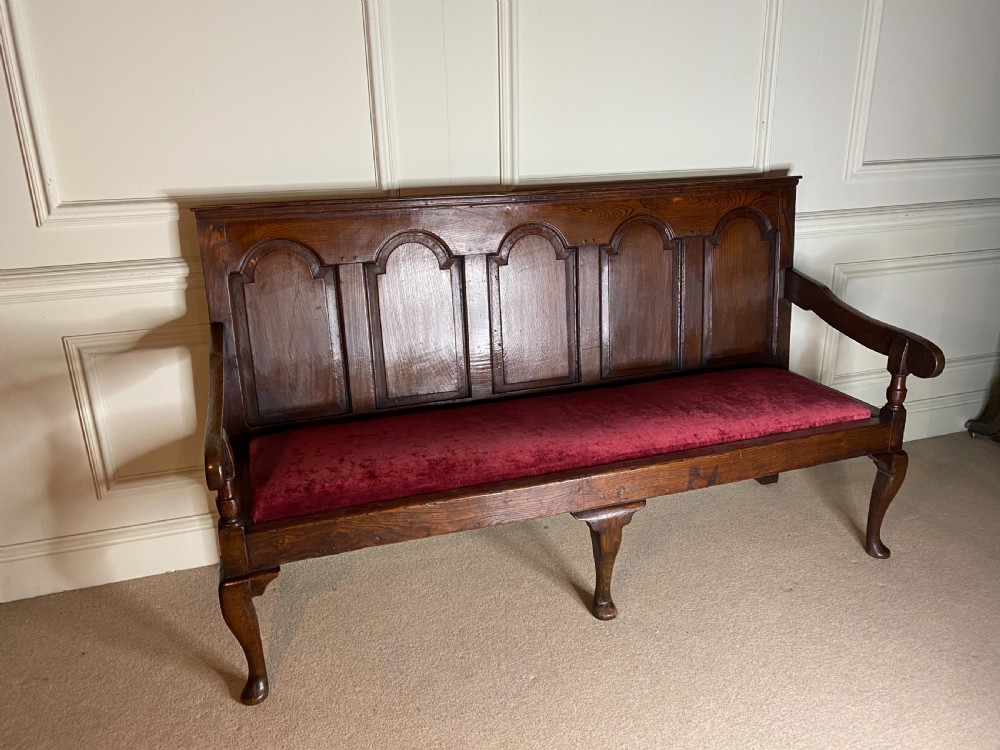 An 18th century dark oak settle featuring a five-panel arched backrest, shaped armrests, cabriole front legs, and a red velvet seat cushion.