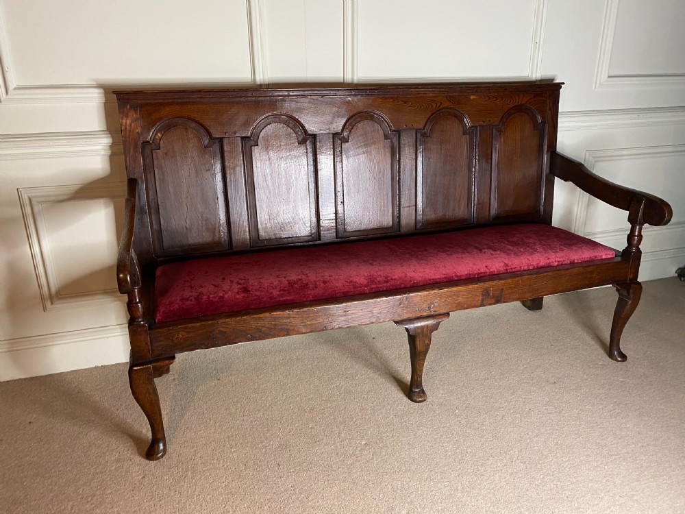 An 18th century dark oak settle featuring a five-panel arched backrest, shaped armrests, cabriole front legs, and a red velvet seat cushion. View II