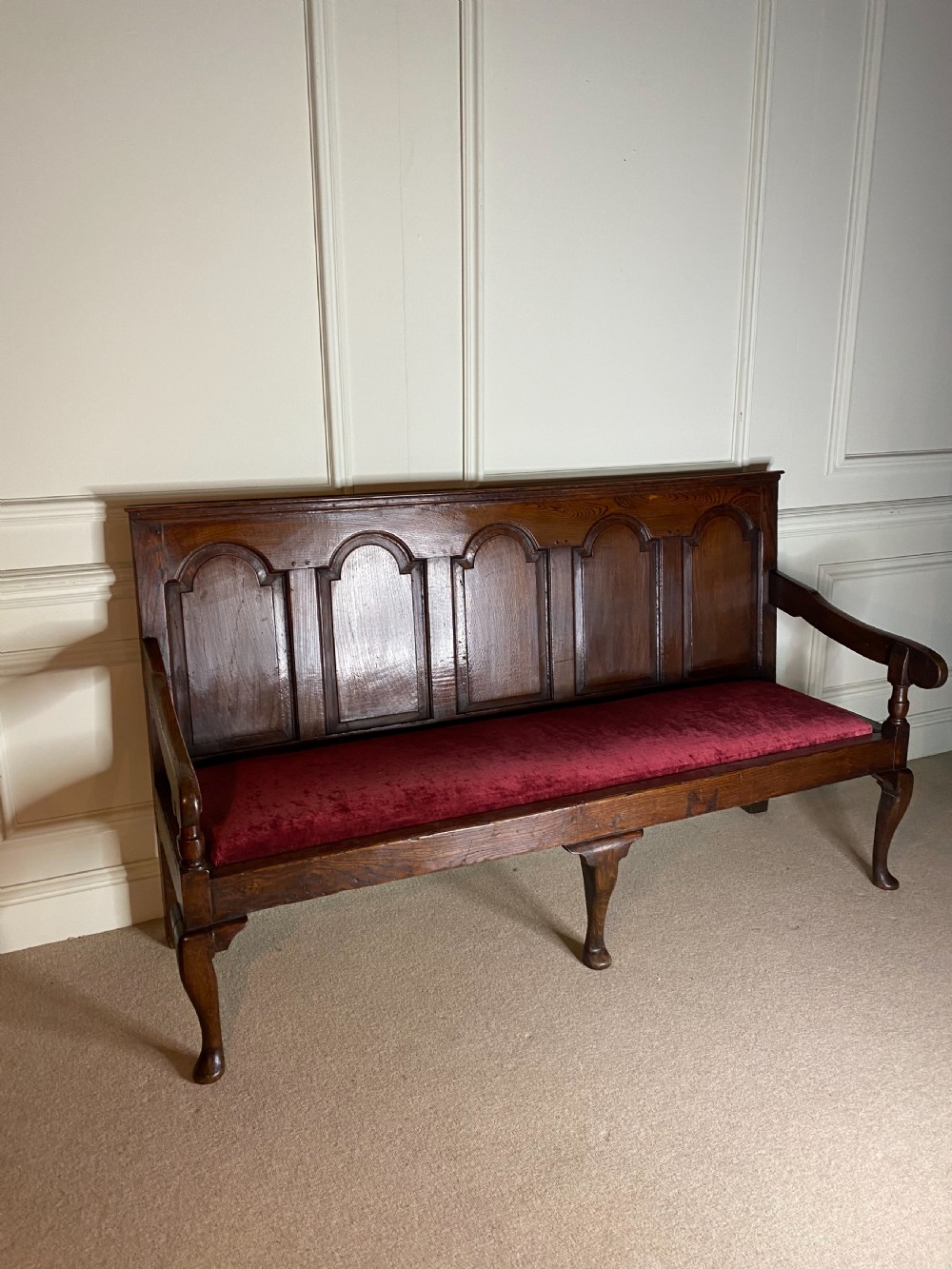 An 18th century dark oak settle featuring a five-panel arched backrest, shaped armrests, cabriole front legs, and a red velvet seat cushion. View III