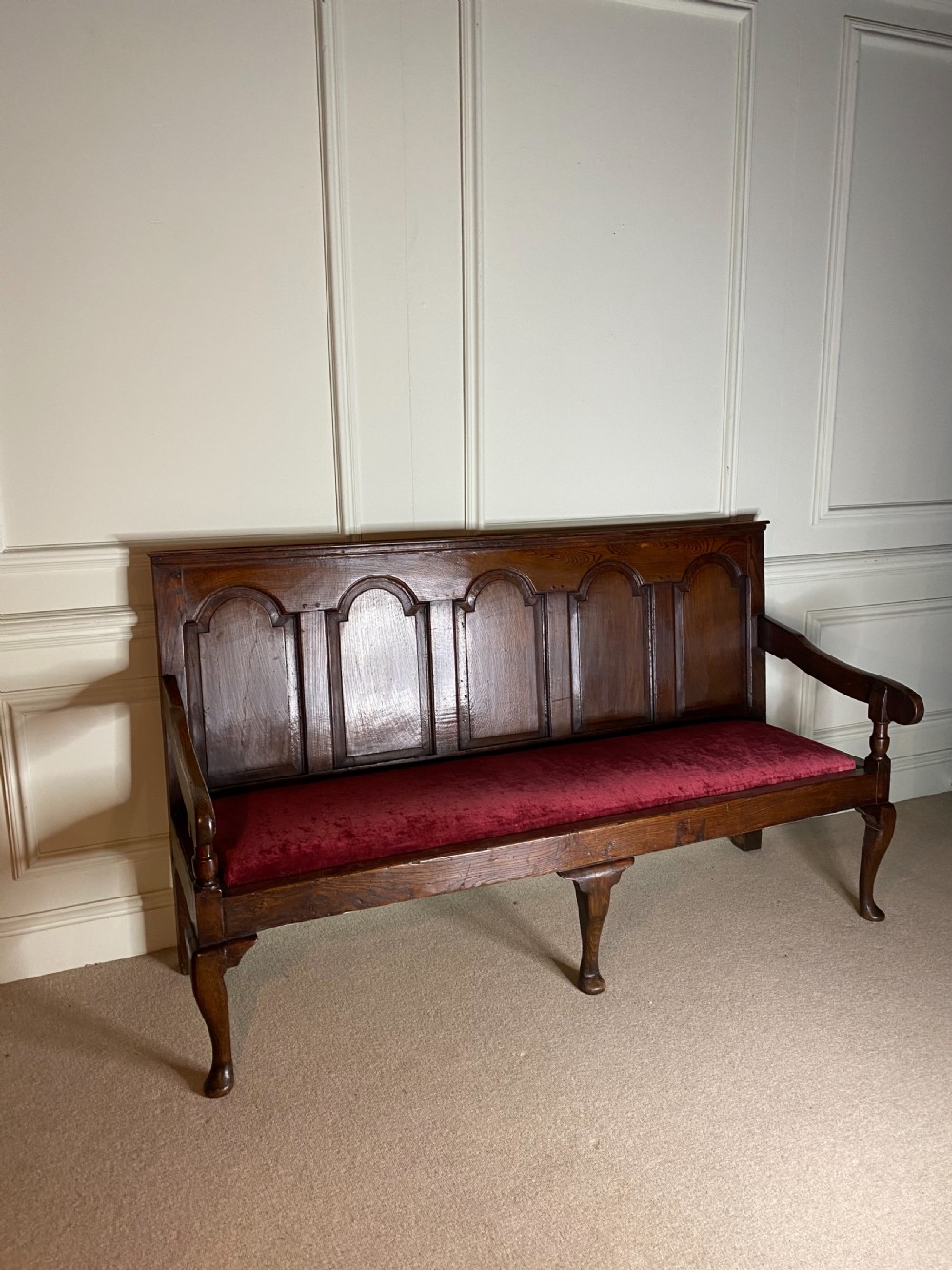 An 18th century dark oak settle featuring a five-panel arched backrest, shaped armrests, cabriole front legs, and a red velvet seat cushion. View V