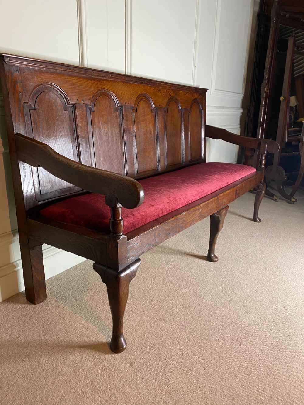 An 18th century dark oak settle featuring a five-panel arched backrest, shaped armrests, cabriole front legs, and a red velvet seat cushion. View VI