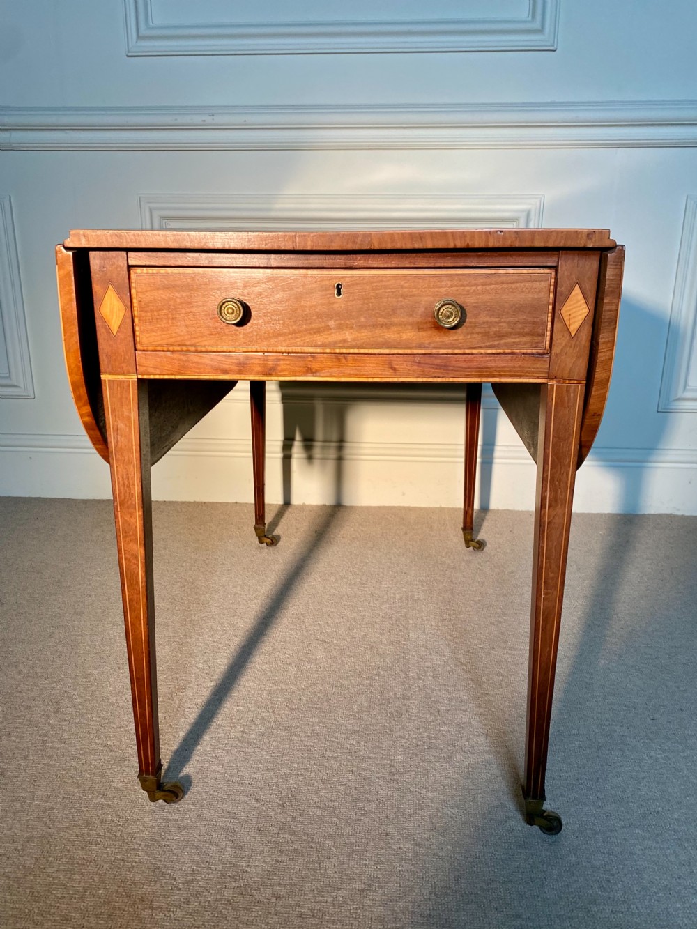 An 18th-century Sheraton mahogany Pembroke table featuring drop leaves, stringing inlay, a single drawer with brass knobs, and square tapered legs on brass castors.