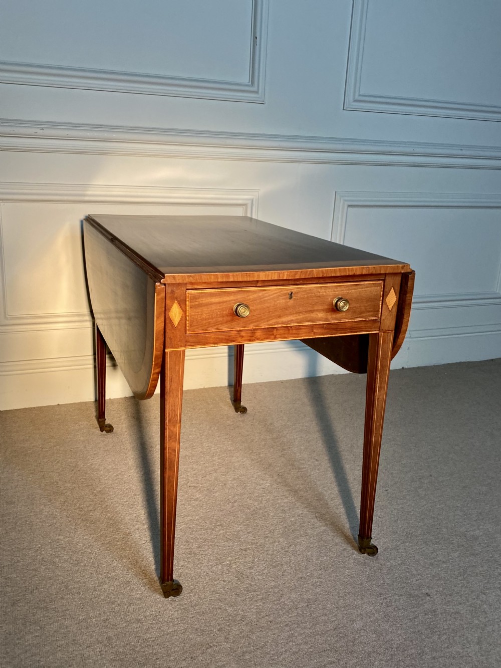 An 18th-century Sheraton mahogany Pembroke table featuring drop leaves, stringing inlay, a single drawer with brass knobs, and square tapered legs on brass castors. View V