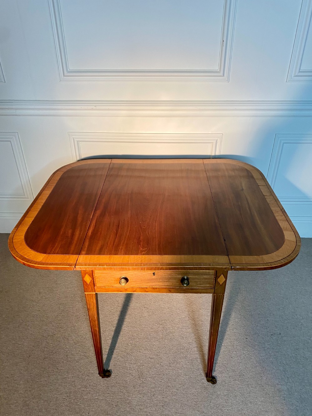 An 18th-century Sheraton mahogany Pembroke table featuring drop leaves, stringing inlay, a single drawer with brass knobs, and square tapered legs on brass castors. View VI