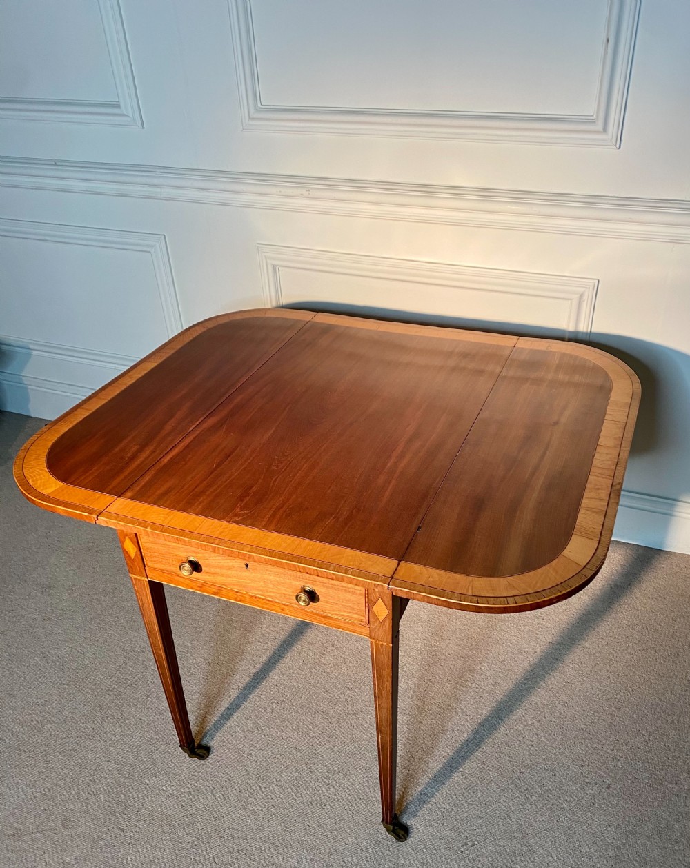 An 18th-century Sheraton mahogany Pembroke table featuring drop leaves, stringing inlay, a single drawer with brass knobs, and square tapered legs on brass castors. View VII