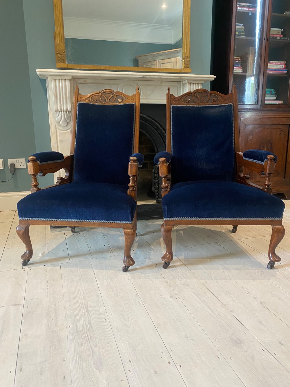 A pair of 19th-century walnut armchairs featuring carved crest rails, padded armrests, brass castors, and deep navy blue velvet upholstery.