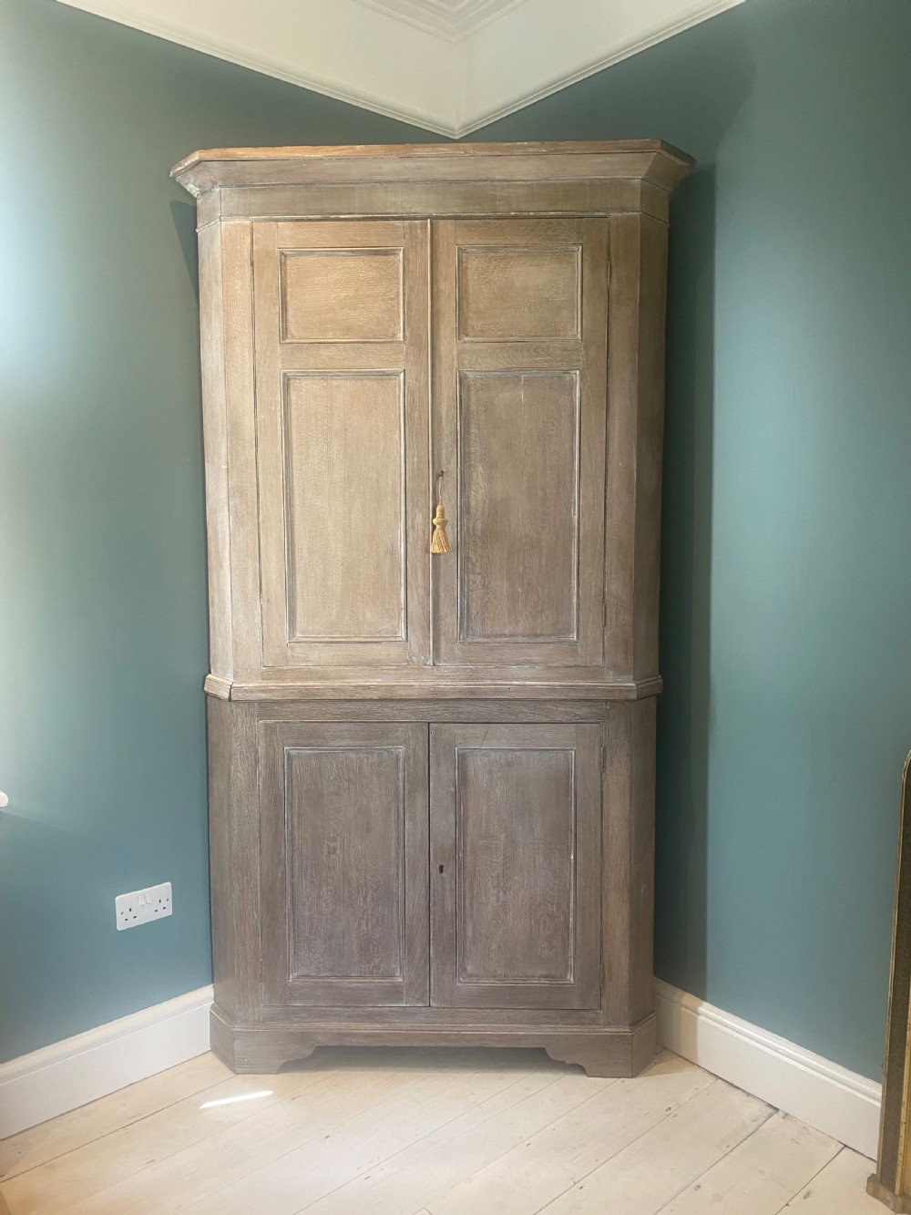An 18th-century two-tier limed oak corner cupboard featuring raised panel doors, a moulded cornice, and a plinth base. View V